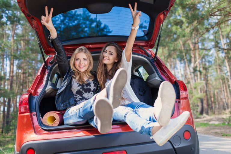 Two college students sit in the open trunk of a red car