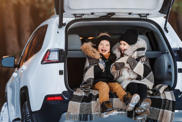 Two children in winter clothes sit in an SUV trunk, wrapped in a plaid blanket, smiling on a snowy day.